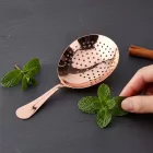 A close-up professional shot of an exclusive copper Julep cocktail strainer, as seen in image_baf12a.png, lying flat on a minimalist dark gray slate surface. Beside the perforated copper strainer are 
