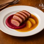 Alaszka fehér porcelán lapostányéron kacsamell céklapürével és narancsmártással, elegáns fine dining tálalásban, meleg természetes fényű éttermi környezetben.