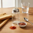 Üveg fűszerszóró chilipelyhekkel és őrölt paprikával, rusztikus konyhai környezetben, textil szalvétával